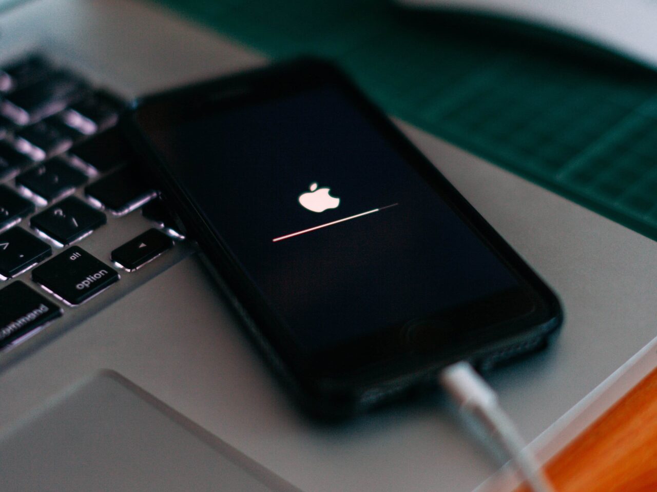 Czarny smartfon leżący na klawiaturze laptopa, wyświetlany jest ekran ładowania systemu z logo Apple. Bateria się ładuje