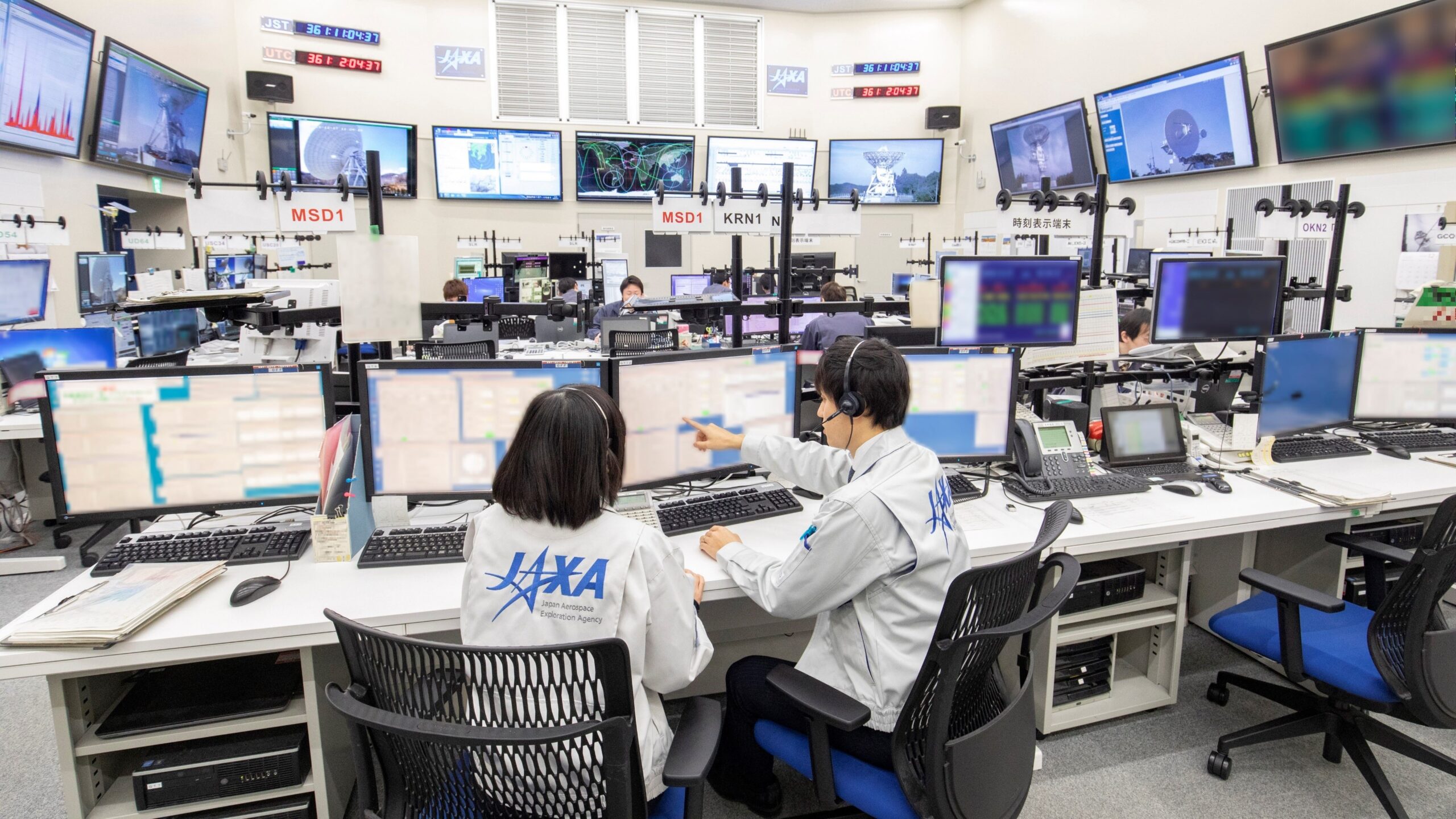 Centrum kontrolne JAXA z pracownikami przy wielu monitorach i sprzęcie komunikacyjnym.