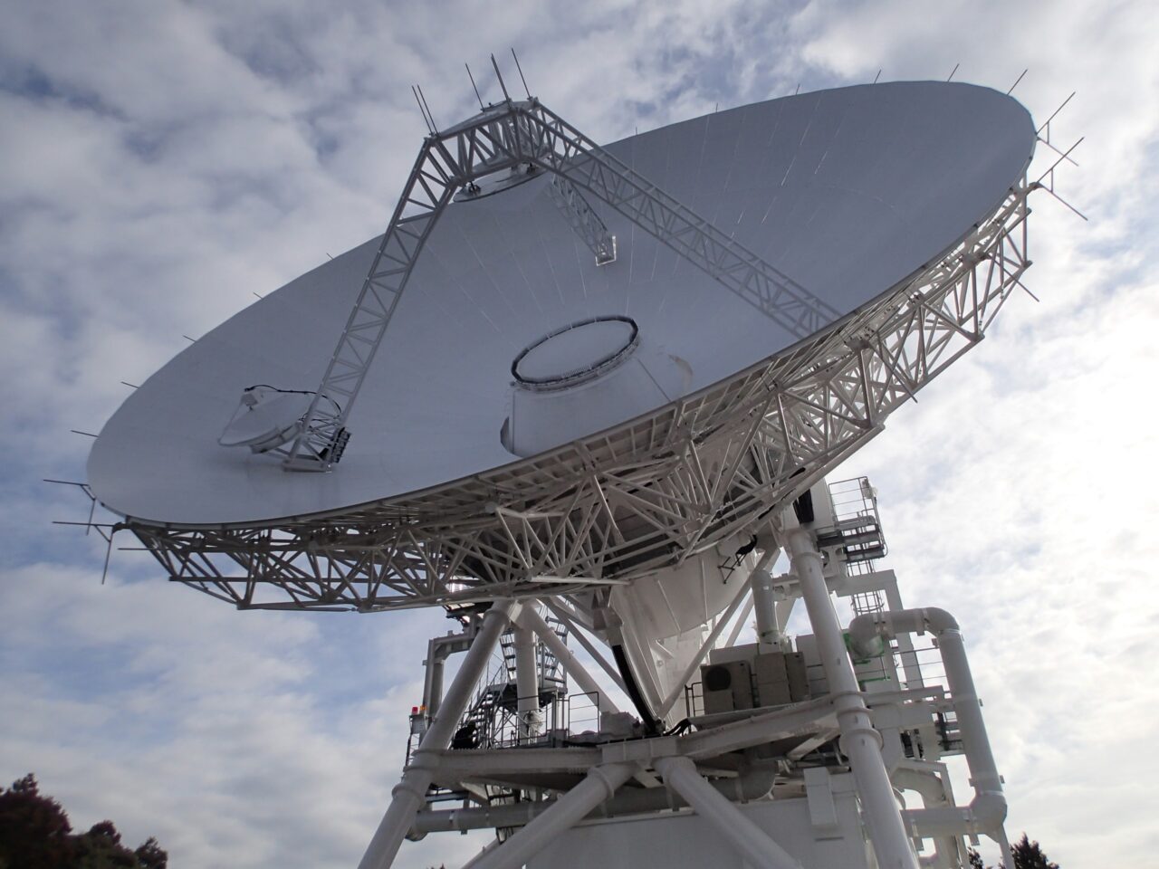 Duża antena satelitarna skierowana ku niebu z zagiętym ramieniem i metalową konstrukcją wsporcza, na tle częściowo zachmurzonego nieba.