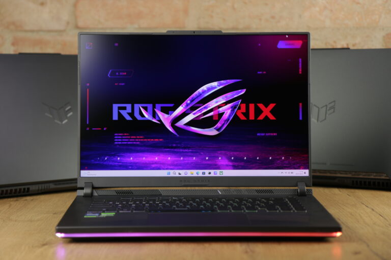 Laptop gamingowy z otwartą pokrywą i wyświetlonym logotypem ROG Strix na ekranie, postawiony na drewnianym biurku, z rozmytym tłem.