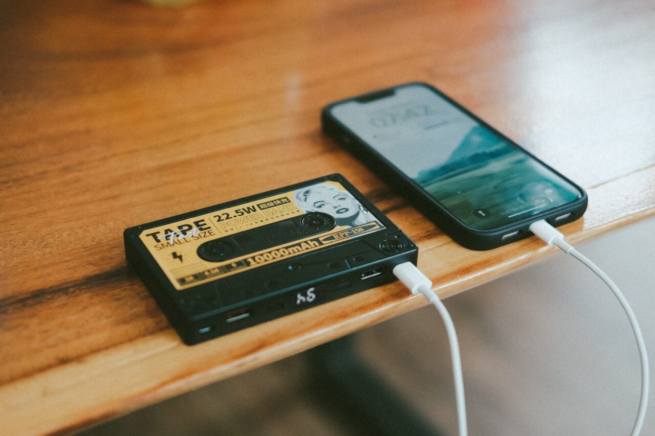 Powerbank w kształcie kasety magnetofonowej podłączony białym kablem do czarnego smartfona leżącego na drewnianym blacie.