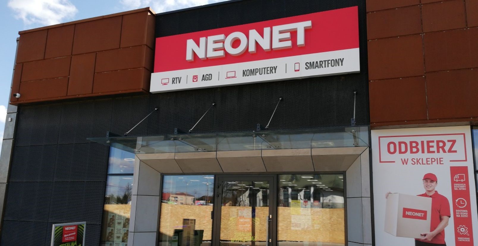 Wejście do sklepu NEONET z witrynami i reklamami, na których widać sprzęt RTV, AGD, komputery i smartfony.