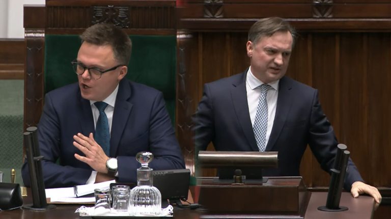 Dwóch mężczyzn w garniturach debatuje za mównicami w izbie parlamentarnej.