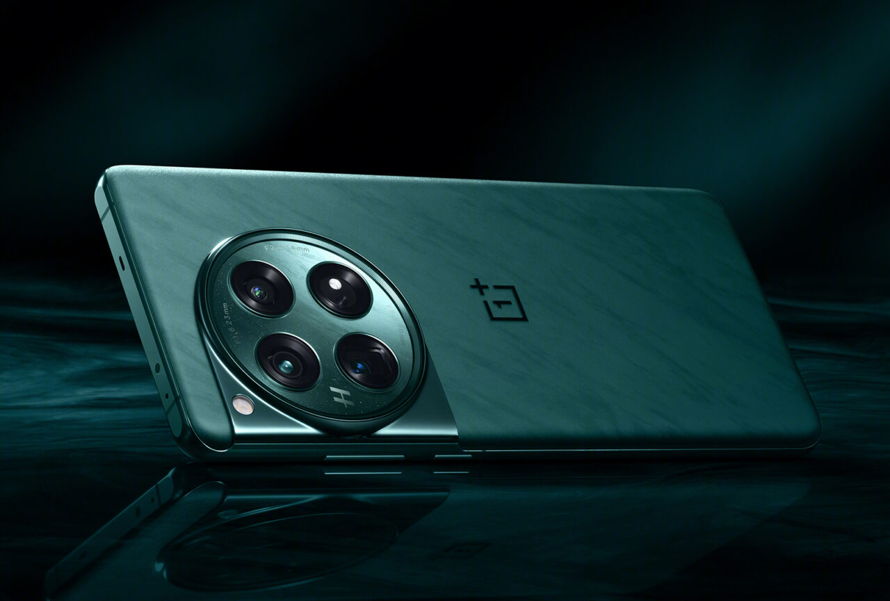 Smartfon OnePlus 12 leżący na boku na ciemnym, odbijającym powierzchni, z układem potrójnych aparatów fotograficznych i logo marki widocznymi z tyłu urządzenia.