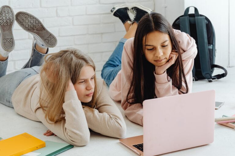 Dwie dziewczynki leżące na podłodze i patrzące na otwarty różowy laptop, z zeszytami i plecakiem w tle.