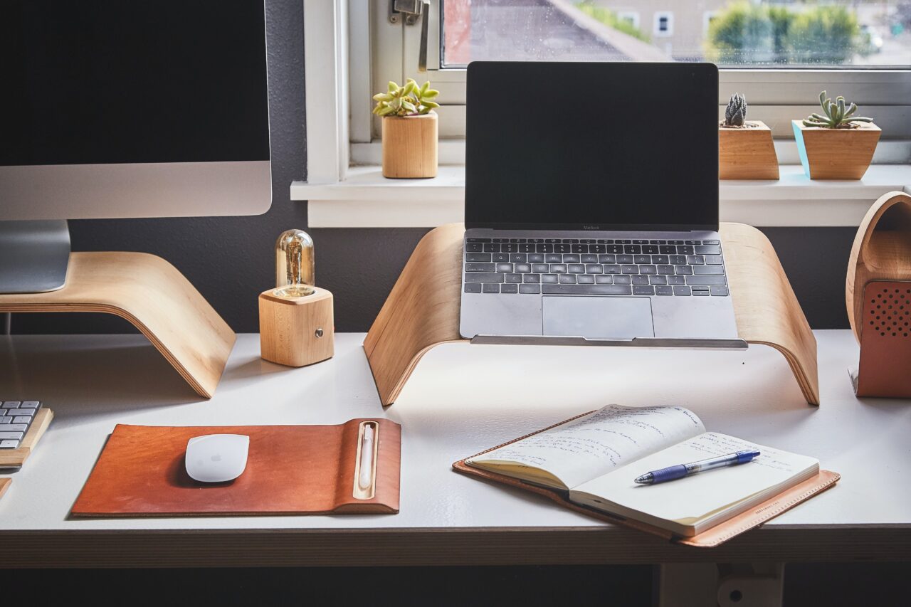 Uma mesa com um laptop sobre um suporte, um caderno, uma caneta, um fragmento de monitor e um mouse.  Uma janela e suculentas ao fundo.