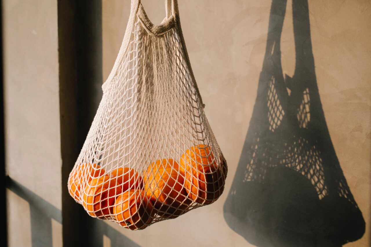 Siatkowa torba na zakupy zawieszona na ścianie zawierająca pomarańcze, z wyraźnym cieniem na tle.