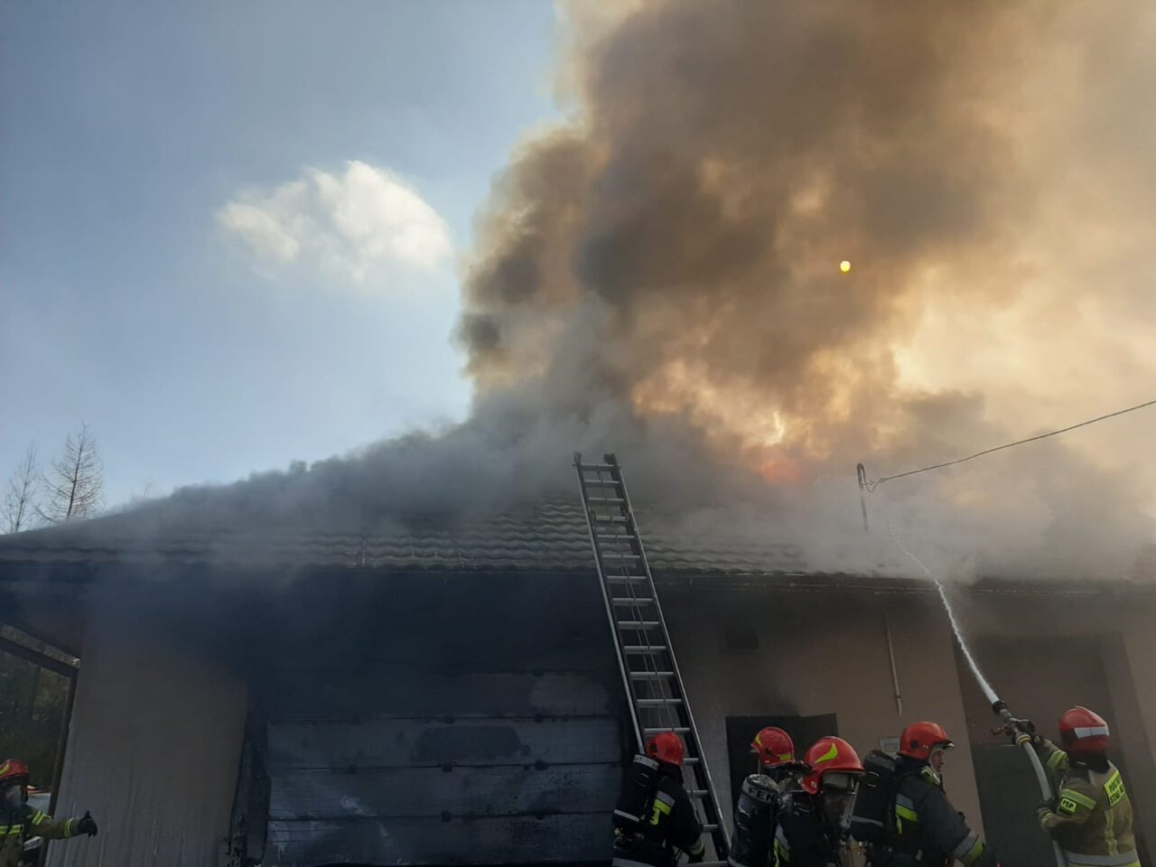 Strażacy gaszą pożar domu, z którego wydobywa się gęsty, ciemny dym. Na zdjęciu widać drabinę opartą o budynek i strumień wody kierowany na płomienie.