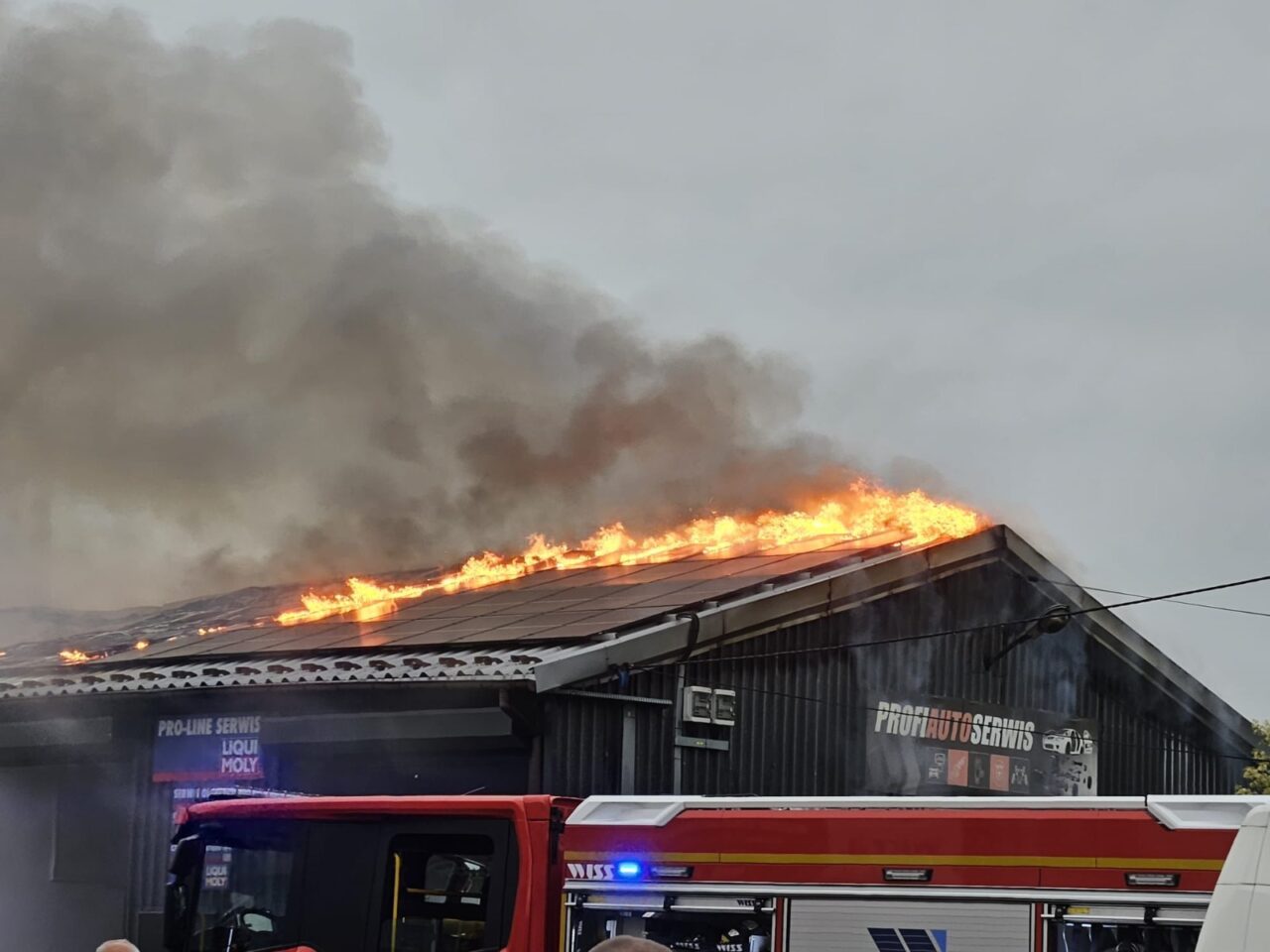 Pożar na dachu budynku przemysłowego z dużą ilością dymu, w tle widać straż pożarną.