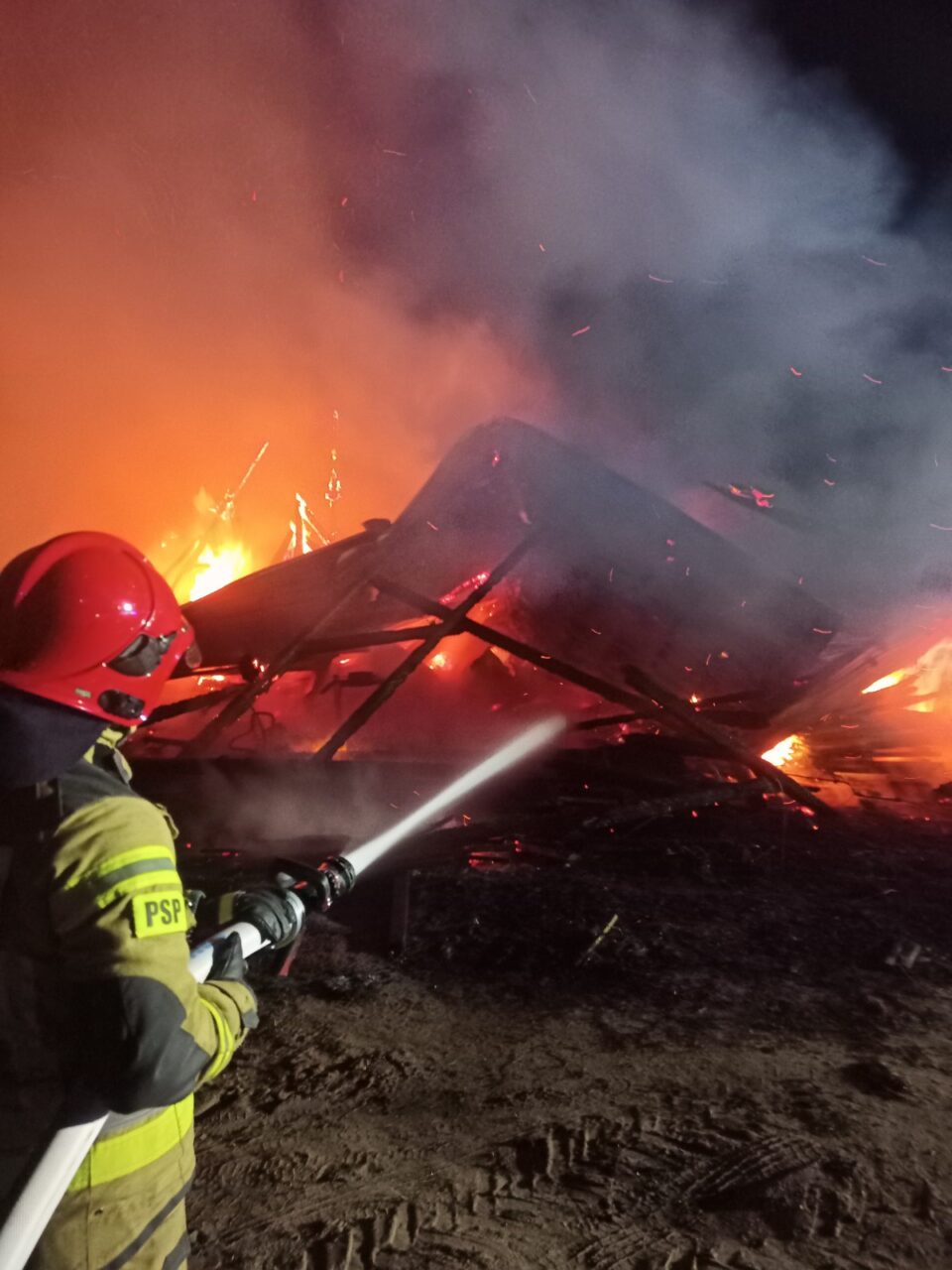 Strażak w pełnym umundurowaniu gasi pożar, trzyma wąż strażacki skierowany w stronę płonących konstrukcji; widoczne są iskry i dym.