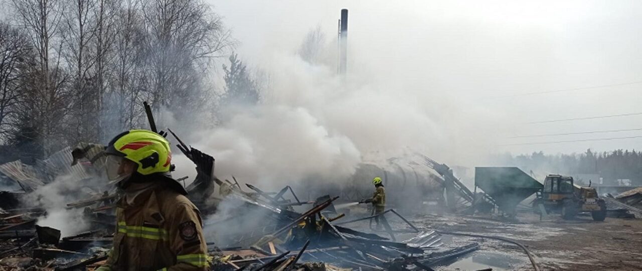 Strażacy w pełnym wyposażeniu gaszą pożar zgliszcz budynku, z którego unosi się gęsty dym; w tle widoczne drzewa i pojazd budowlany. Pożary fotowoltaiki w Polsce