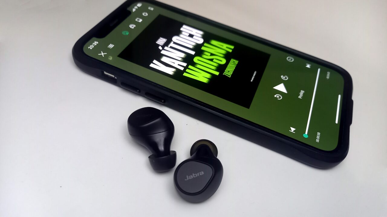 Smartfon z ekranem wyświetlającym audiobooka oraz obok niego bezprzewodowe słuchawki marki Jabra.