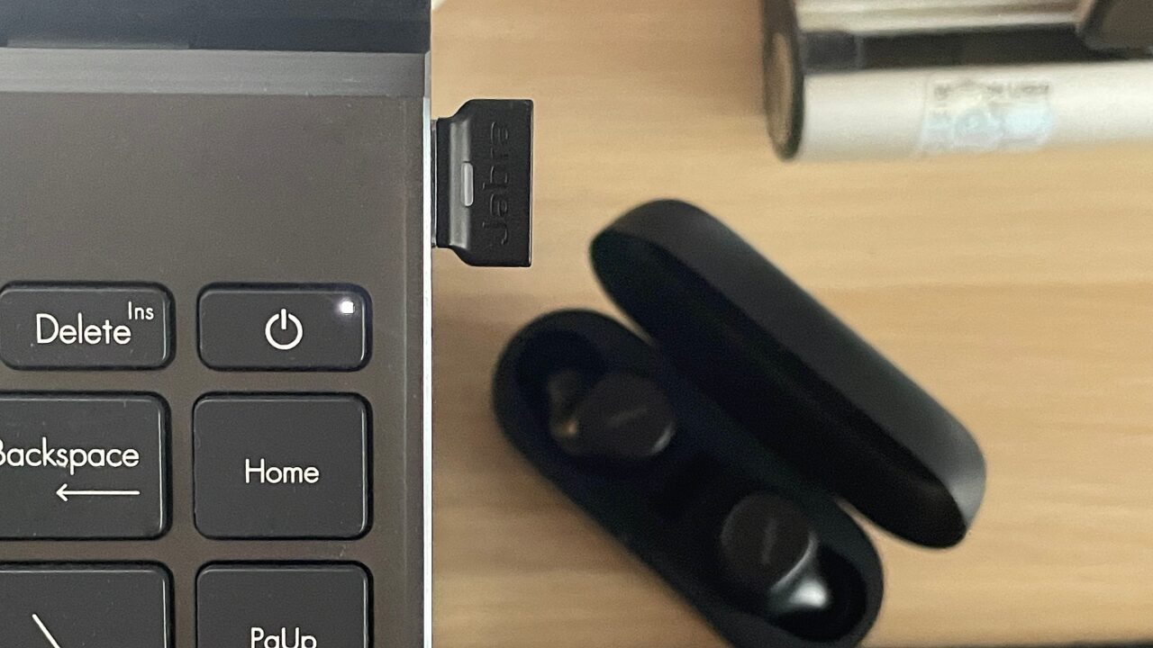Część laptopa z klawiszami "Delete" i "Power" obok otwartego portu USB, w którym znajduje się czarny pendrive, oraz czarne bezprzewodowe słuchawki w otwartym etui na biurku.