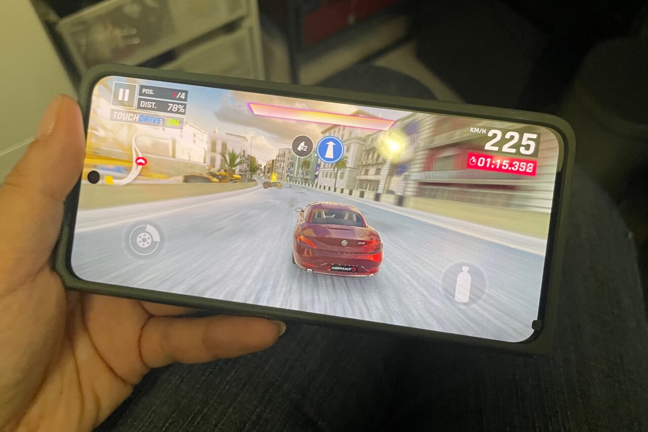 Osoba trzyma smartfona, na którym uruchomiona jest gra wyścigowa z czerwonym samochodem na asfaltowym torze w miejskim otoczeniu. Na ekranie wyświetlane są: prędkość 225 km/h, czas okrążenia oraz interfejs użytkownika gry.