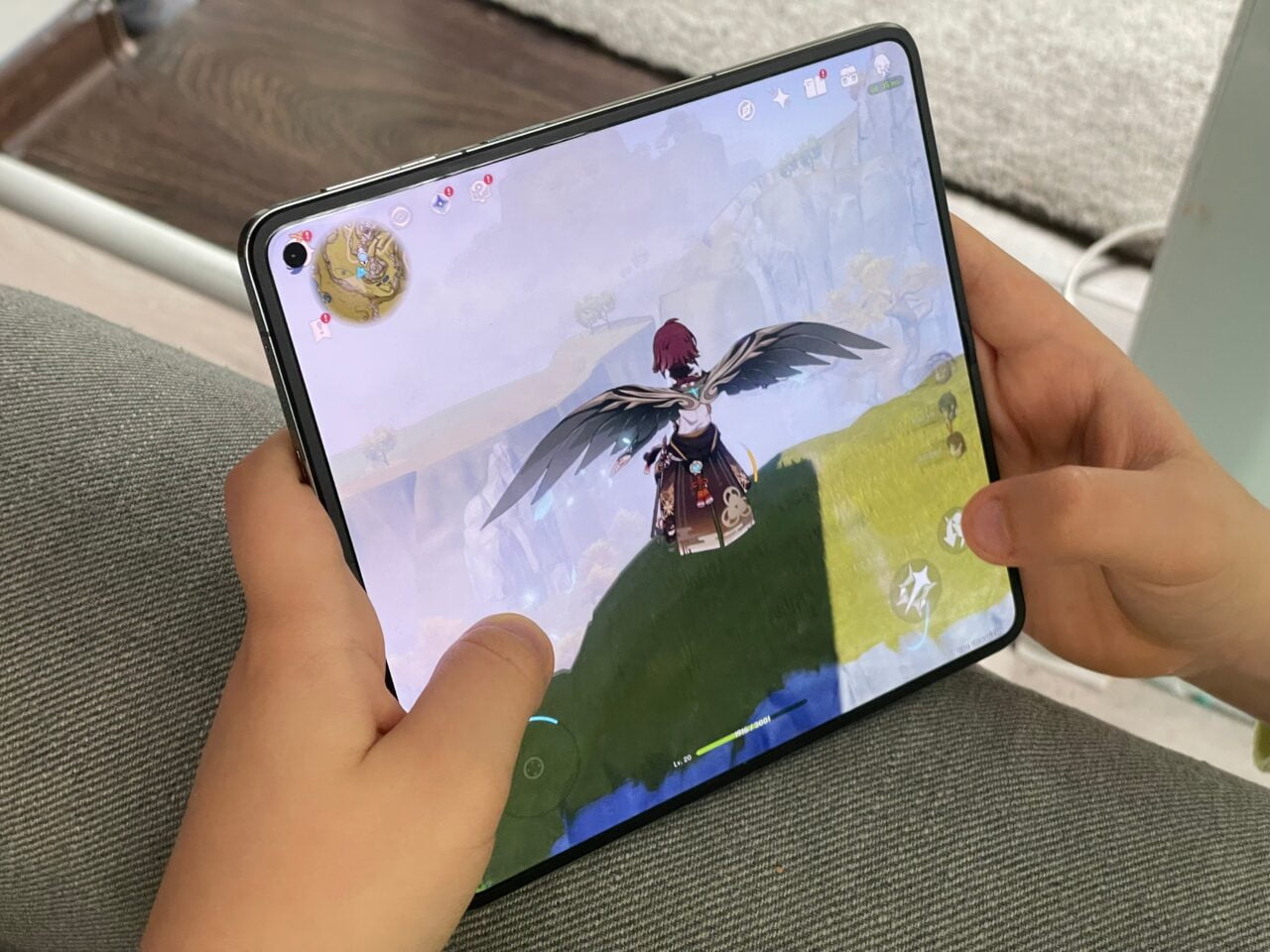 Dziecko trzyma tablet z wyświetlaną grą, na ekranie postać z czarnymi skrzydłami przelatuje nad zielonym terenem w wirtualnym świecie.  recenzja oneplus open, genshin impact