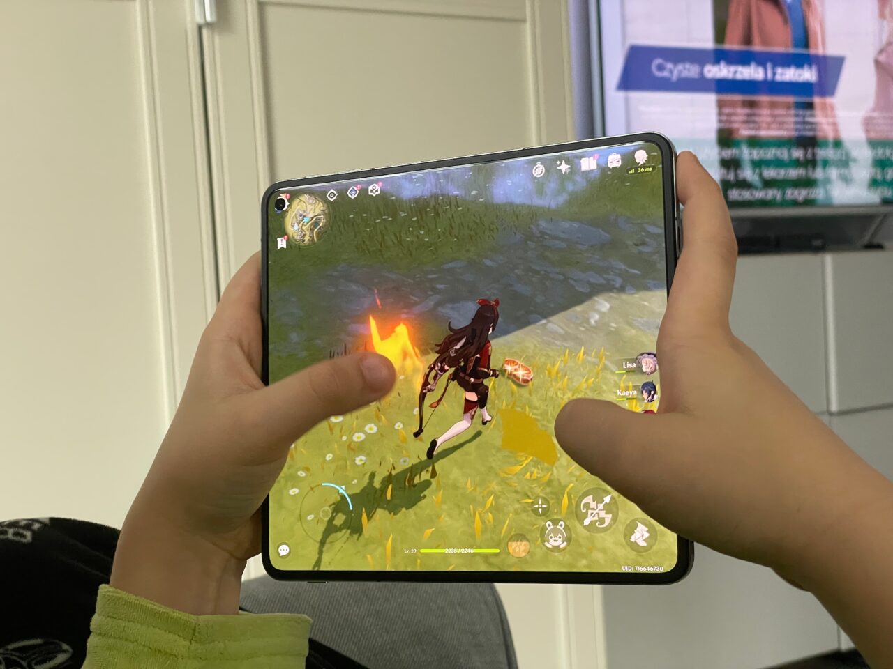Osoba trzymająca tablet, na którym wyświetlana jest gra z postacią biegnącą przez trawiaste pole z widocznym ogniem w grze mobilnej, w tle nieostry ekran telewizora. recenzja oneplus open, genshin impact