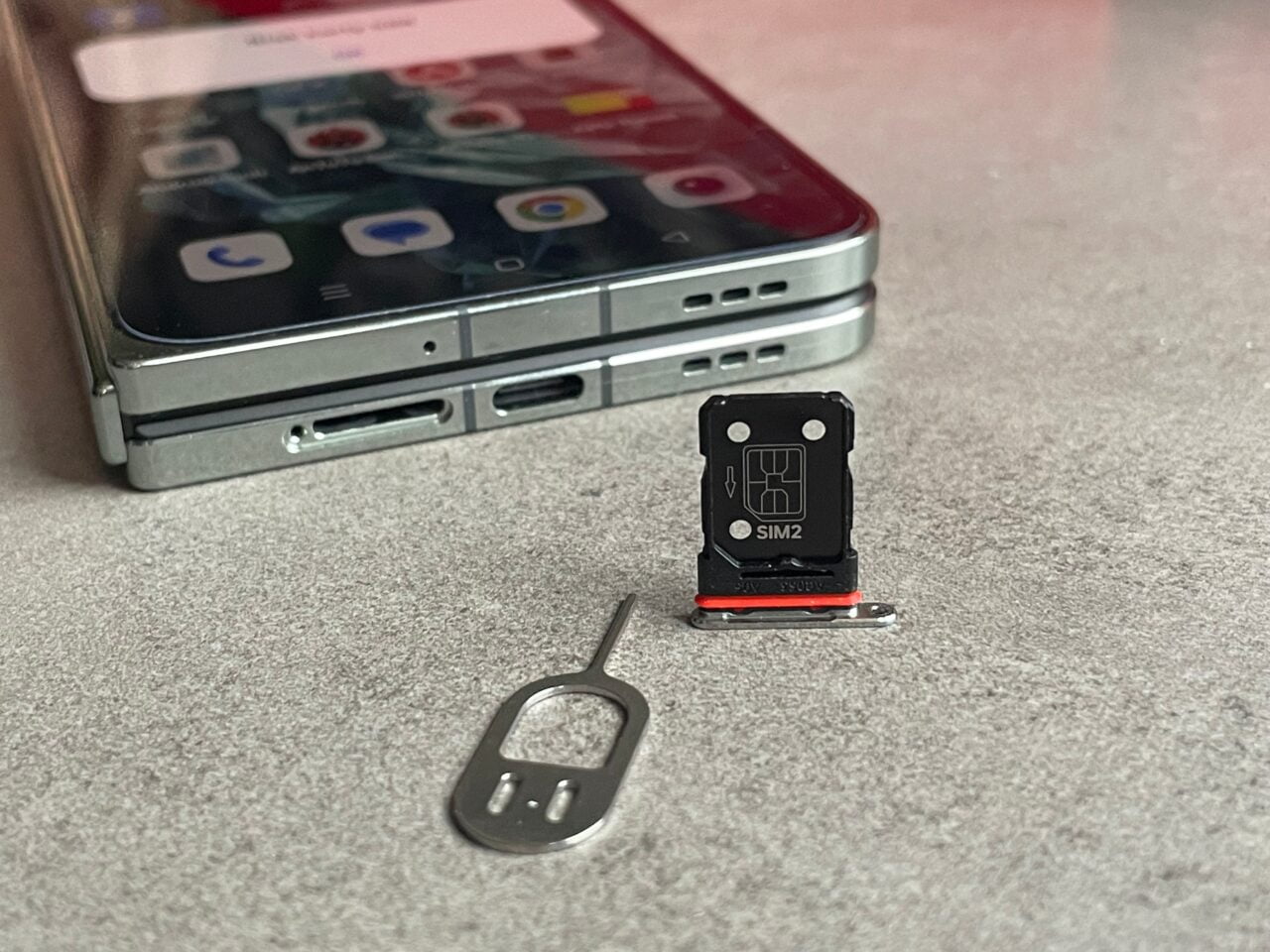 Smartfon z otwartym tacką na karty SIM obok wyciągniętej tacki z oznaczeniem SIM2 oraz narzędziem do jej wyjęcia na szarym tle. recenzja oneplus open