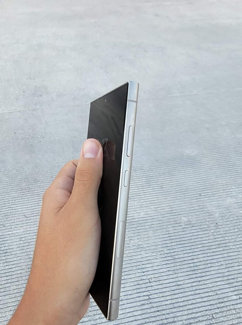 Częściowo widoczna dłoń trzymająca smartfon samsung galaxy s24 ultra w pozycji bocznej na tle betonowej powierzchni.