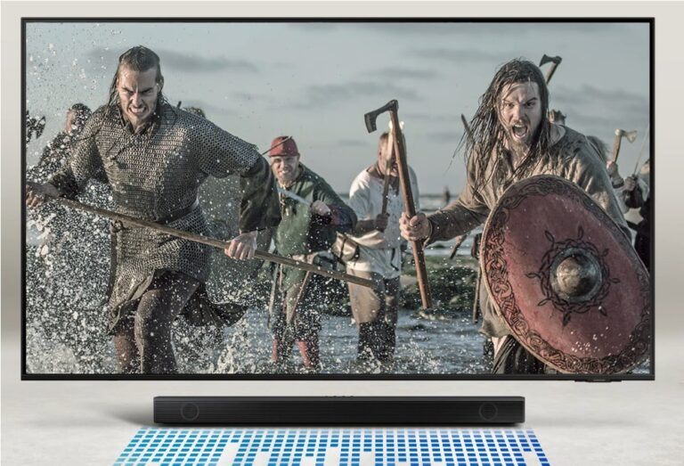 Scena przedstawiająca wojowników wikingów brodzących przez płytką wodę, wyposażonych w zbroje i trzymających broń, na ekranie telewizora znajdującego się nad soundbarem