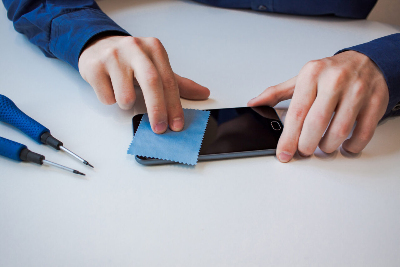 Osoba czyści ekran smartfona za pomocą niebieskiej ściereczki, obok leżą śrubokręty.