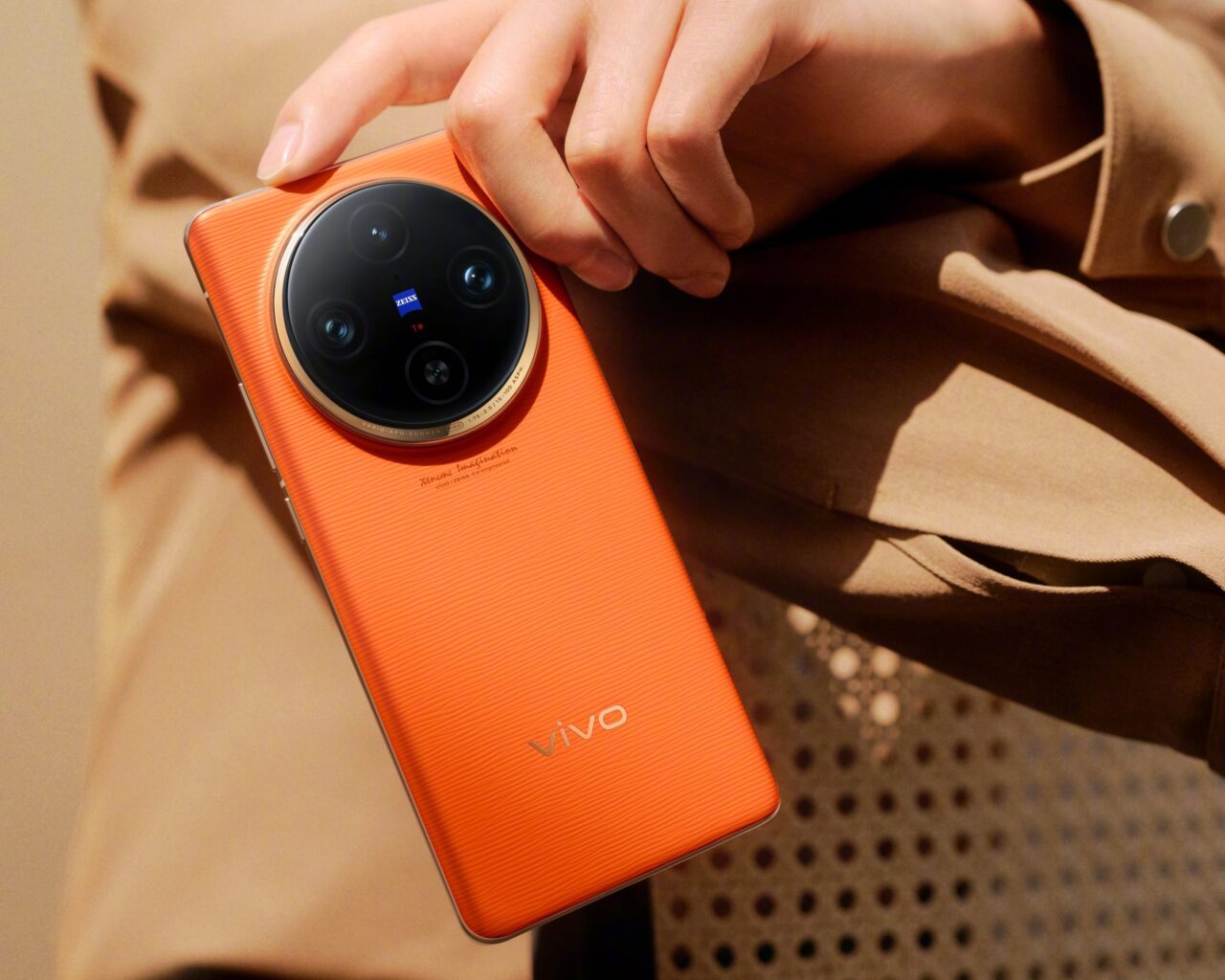 Pomarańczowy smartfon vivo x100 z procesorem Dimensity 9300 trzymany w ręku na beżowym tle.