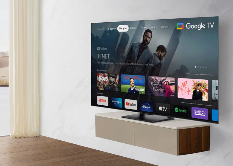 Telewizor z płaskim ekranem wyświetlający interfejs Google TV umieszczony na niskiej białej szafce w jasnym pomieszczeniu z drewnianą podłogą.