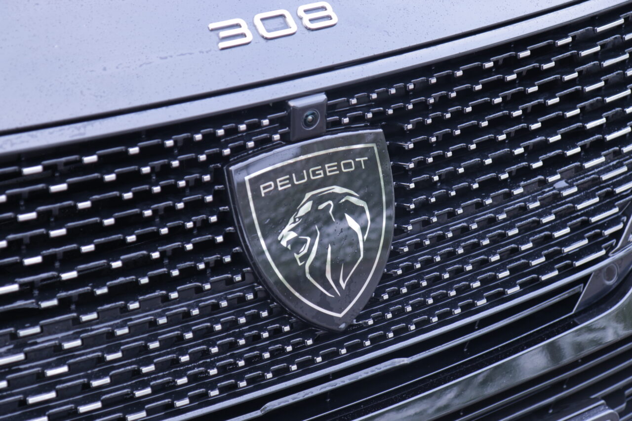 Zbliżenie na logo testowanego Peugeot 308 III z umieszczonym symbolem lwa na czarnej atrapie chłodnicy, nad którym widoczne są srebrne cyfry "308".