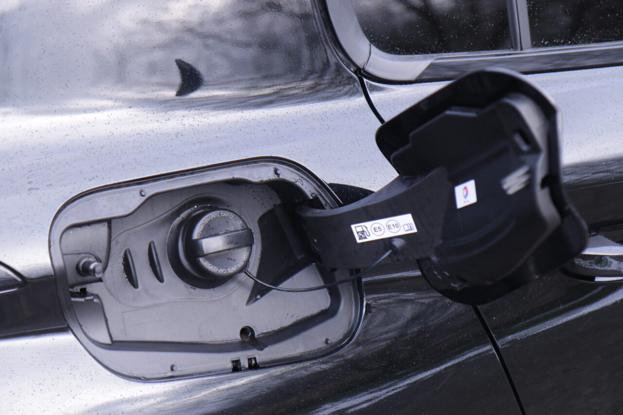 Zdjęcie otwartego wlewu paliwa samochodowego z korkiem przytwierdzonym paskiem bezpieczeństwa, pokazujące instrukcje dotyczące rodzajów paliw, z widocznym symbolem paliwa z domieszką etanolu E5 i E10. Test Peugeot 308 III
