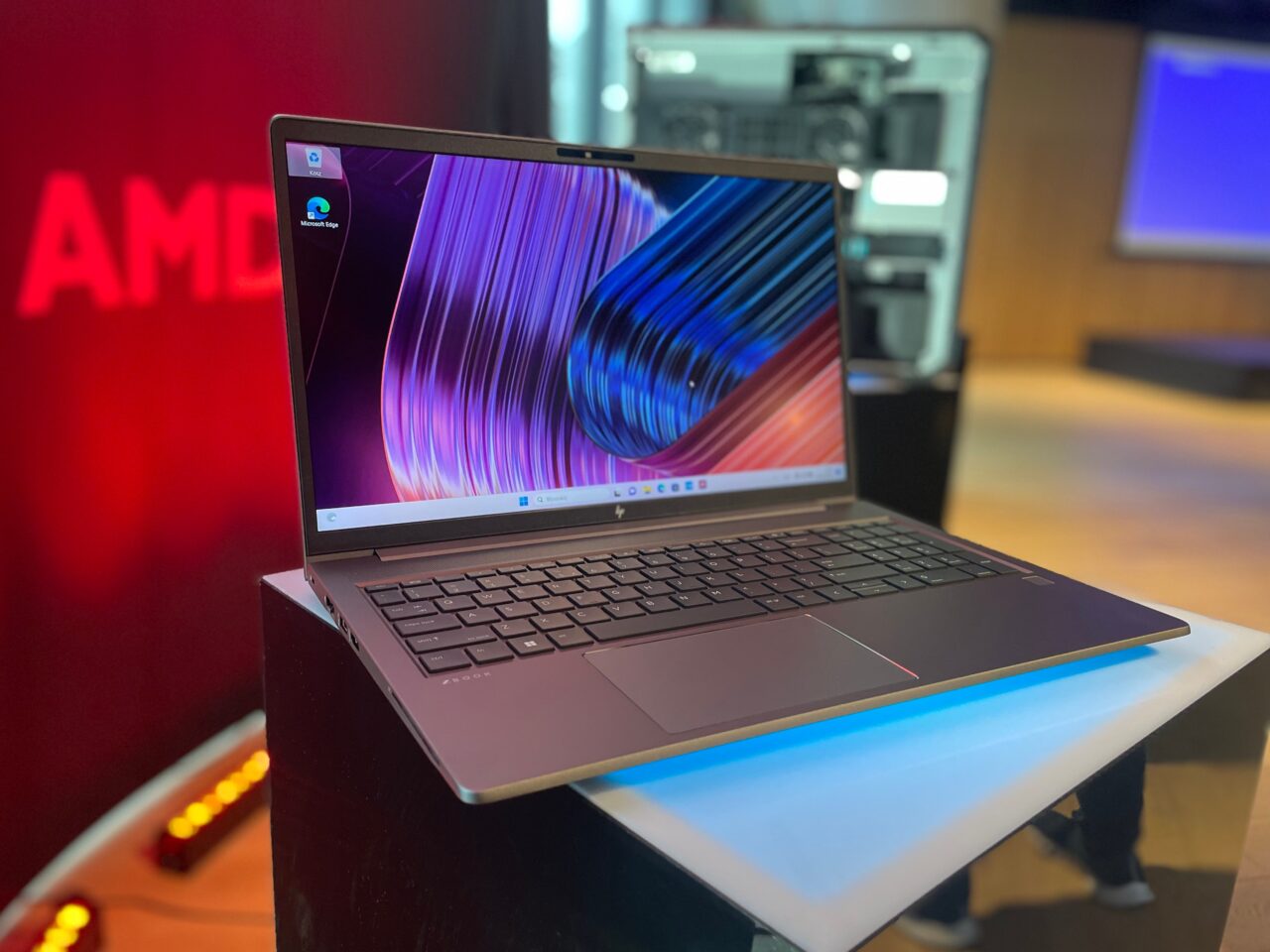 Laptop HP na stole z włączonym ekranem prezentującym kolorową tapetę, w tle rozmyty napis AMD.