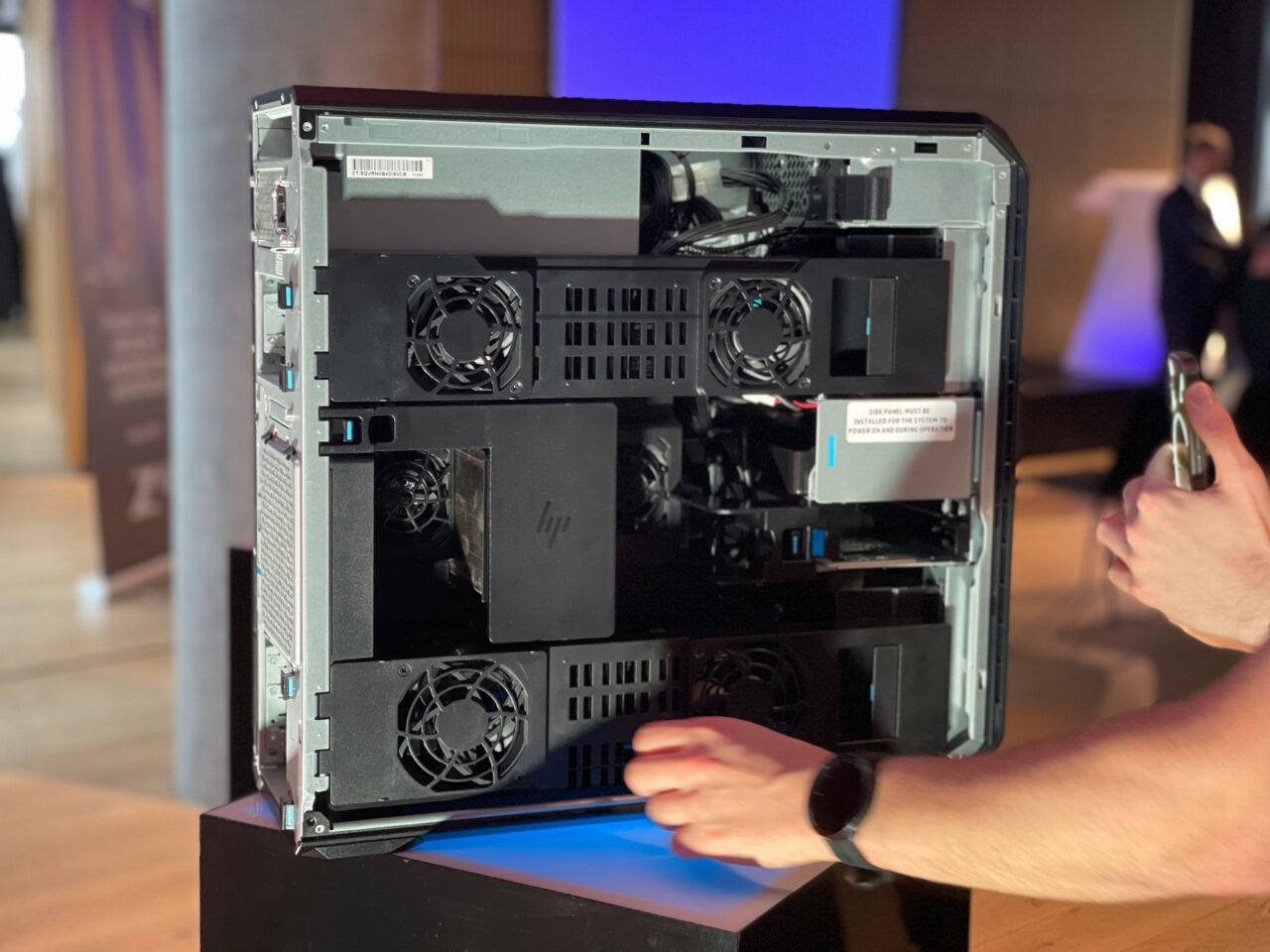Obudowa komputera stacjonarnego HP z otwartym panelem bocznym pokazującym wewnętrzne komponenty, w tym karty graficzne i wentylatory chłodzące. Obracana jest przez osobę znajdującą się po prawej stronie zdjęcia.