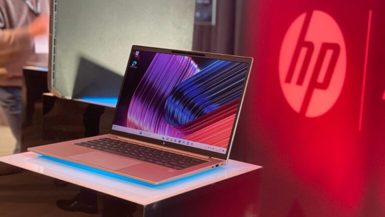 Laptop marki HP na wystawie z otwartą pokrywą i włączonym ekranem pokazującym kolorową tapetę, w tle czerwone logo HP i rozmazana postać człowieka.