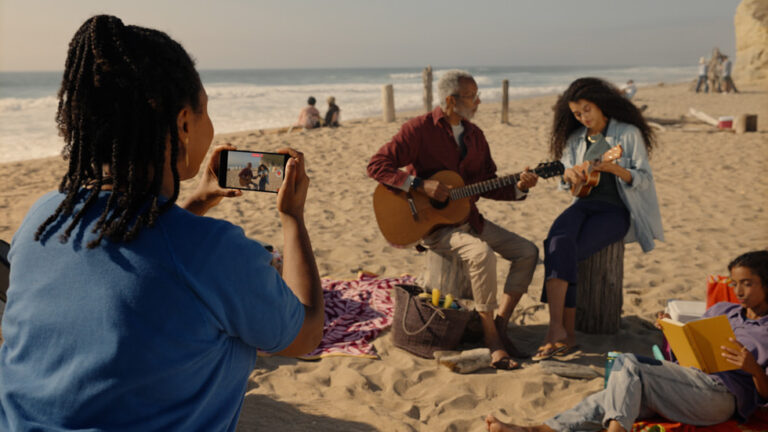 Osoba nagrywa wideo przestrzenne Apple iPhone na plaży dwóm grającym na gitarach ludziom, obok dziecko czyta książkę.