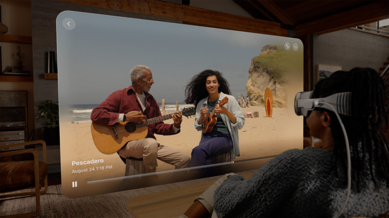 Osoba nosząca zestaw gogli VR siedzi w pomieszczeniu, a na ekranie wyświetlana jest wirtualna rzeczywistość przedstawiająca dwie grające na gitarach osoby na plaży.