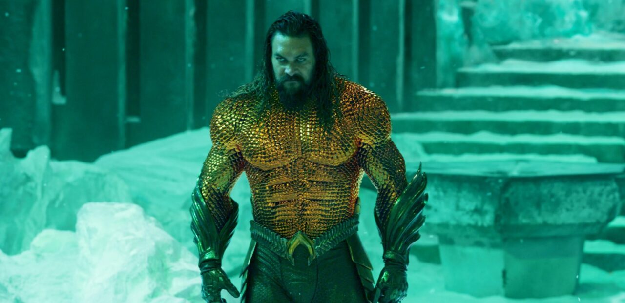 Mężczyzna w złotej zbroi inspirowanej łuskami ryb, z długimi czarnymi włosami, stoi w zielonkawo oświetlonym pomieszczeniu z lodowymi elementami i kamiennymi schodami w tle. Kadr z filmu Aquaman i Zaginione Królestwo