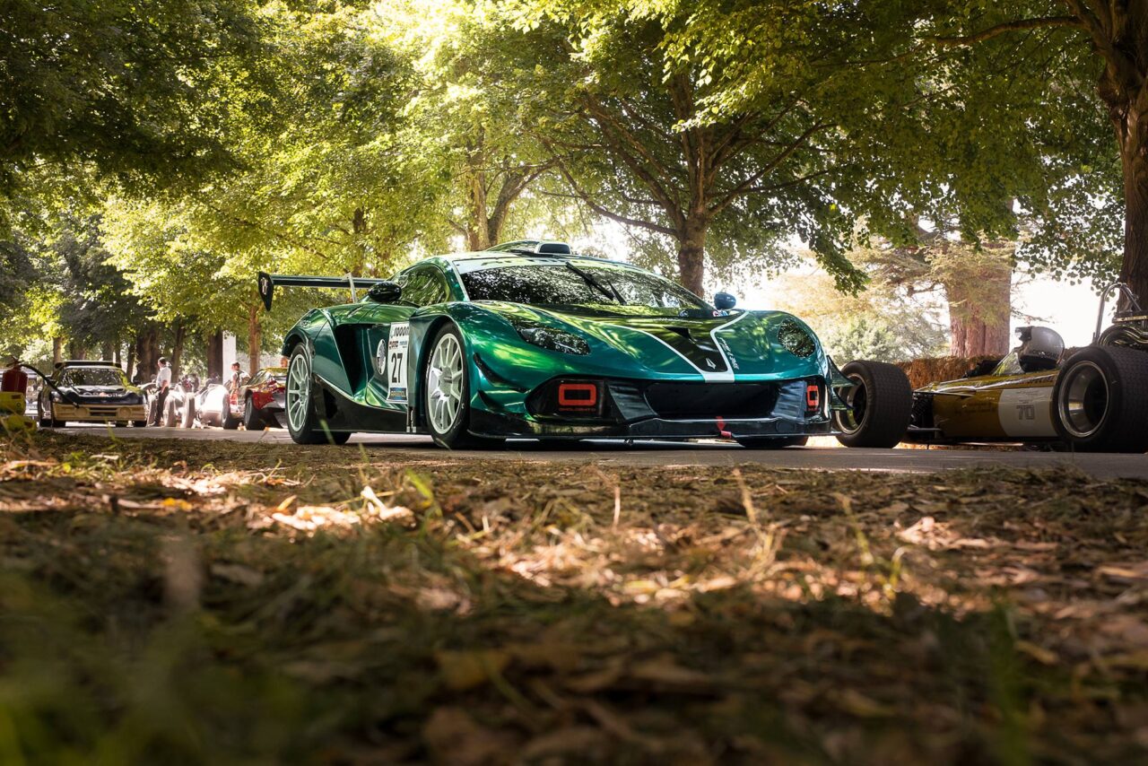 Zielony samochód sportowy Arrinera Hussarya z dużym spoilerem na tle drzew i innych klasycznych samochodów wyścigowych przy słonecznym dniu.