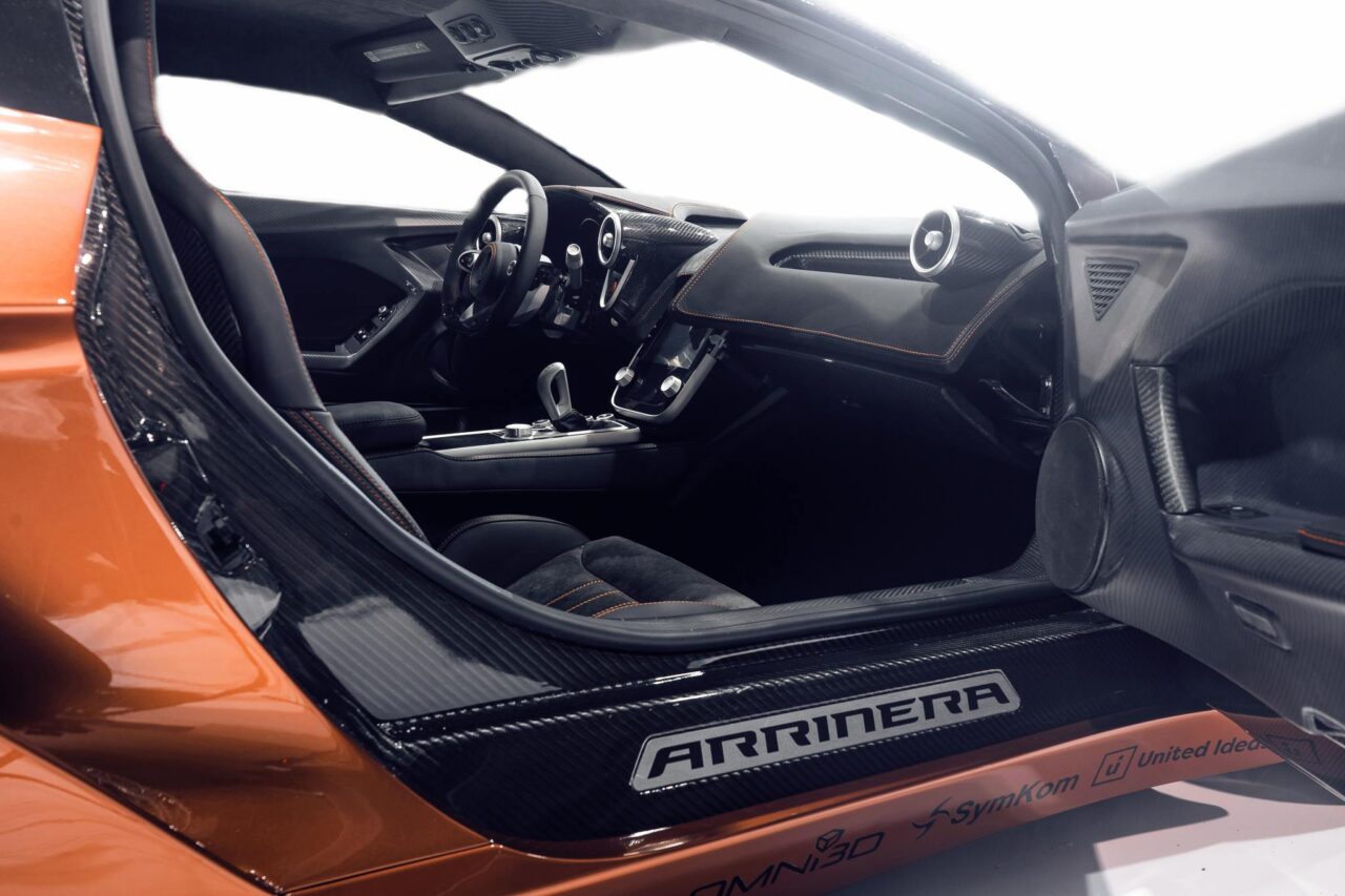 Wnętrze sportowego samochodu marki Arrinera Hussarya, widok na kierownicę i deske rozdzielczą, akcenty z włókna węglowego, podświetlenie, pomarańczowe i czarne wykończenie.