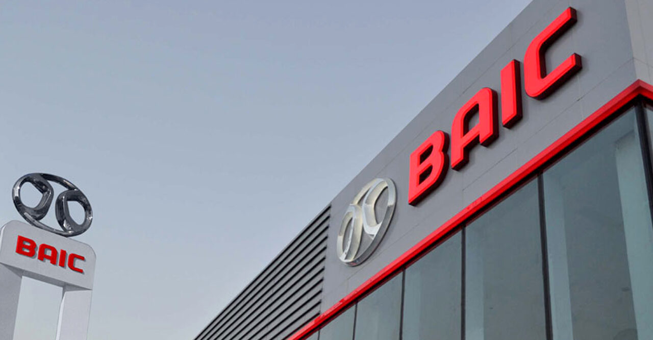 Widok na budynek z logo BAIC podświetlonym na czerwono na elewacji oraz srebrne, metaliczne logo BAIC na wolnostojącej tablicy.