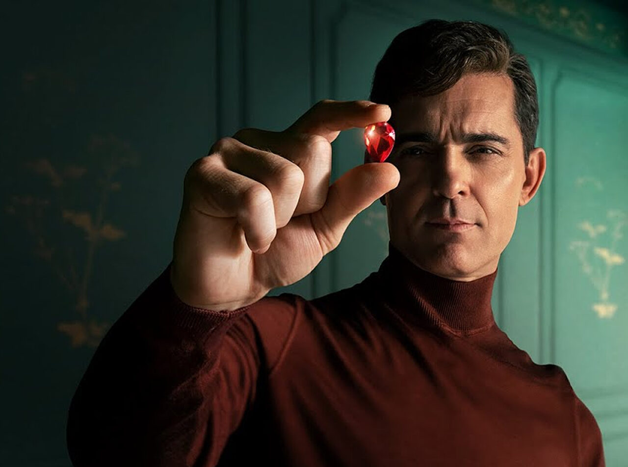 Mężczyzna w bordowym golfie trzyma czerwony, błyszczący obiekt między palcami. Główny bohater serialu Berlin będącego jedną z nowości na Netflix