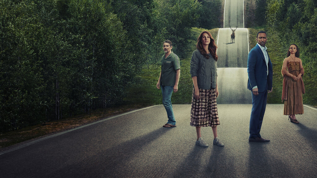 Cztery osoby stoją na asfaltowej drodze, otoczonej przez zielone drzewa, zwrócone w stronę obserwatora. Od lewej: mężczyzna w zielonej koszuli i jeansach, kobieta w szarej kurtce i wzorzystej spódnicy, mężczyzna w ciemnoniebieskim garniturze oraz kobieta w brązowej sukience. W tle na środku drogi widać jelenia.