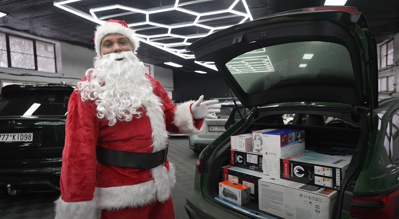 Mężczyzna przebrany za Świętego Mikołaja stoi obok otwartego bagażnika zielonego samochodu, w którym znajdują się zapakowane prezenty, w tym pudełka z elektroniką.