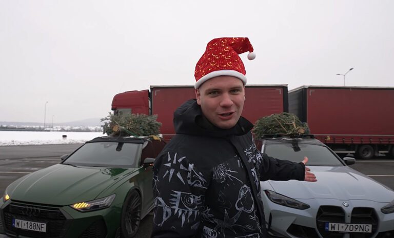 Mężczyzna (Budda) w świątecznej czapce stojący na parkingu z ciężarówkami, pokazujący na zielony i srebrny samochód osobowy z choinką przewiązaną na dachu.