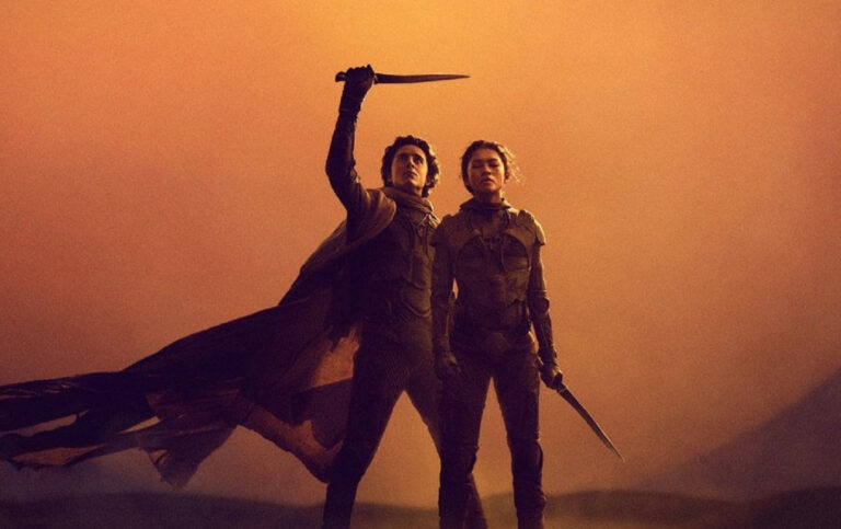 Dwóch bohaterów w futurystycznych strojach stojących na pustyni z unoszącym się pyłem, mężczyzna trzyma miecz w górze, kolorystyka zdjęcia w ciepłych, pomarańczowych tonach. Zdjęcie z Diuna 2 ilustrujące artykuł pt. zapowiedzi filmowe na 2024 rok