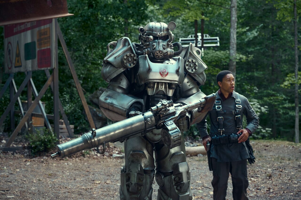 Uma figura em uma armadura futurista com armas que lembram uma minigun está ao lado de um homem com roupas táticas, com um ambiente de floresta ao fundo.
