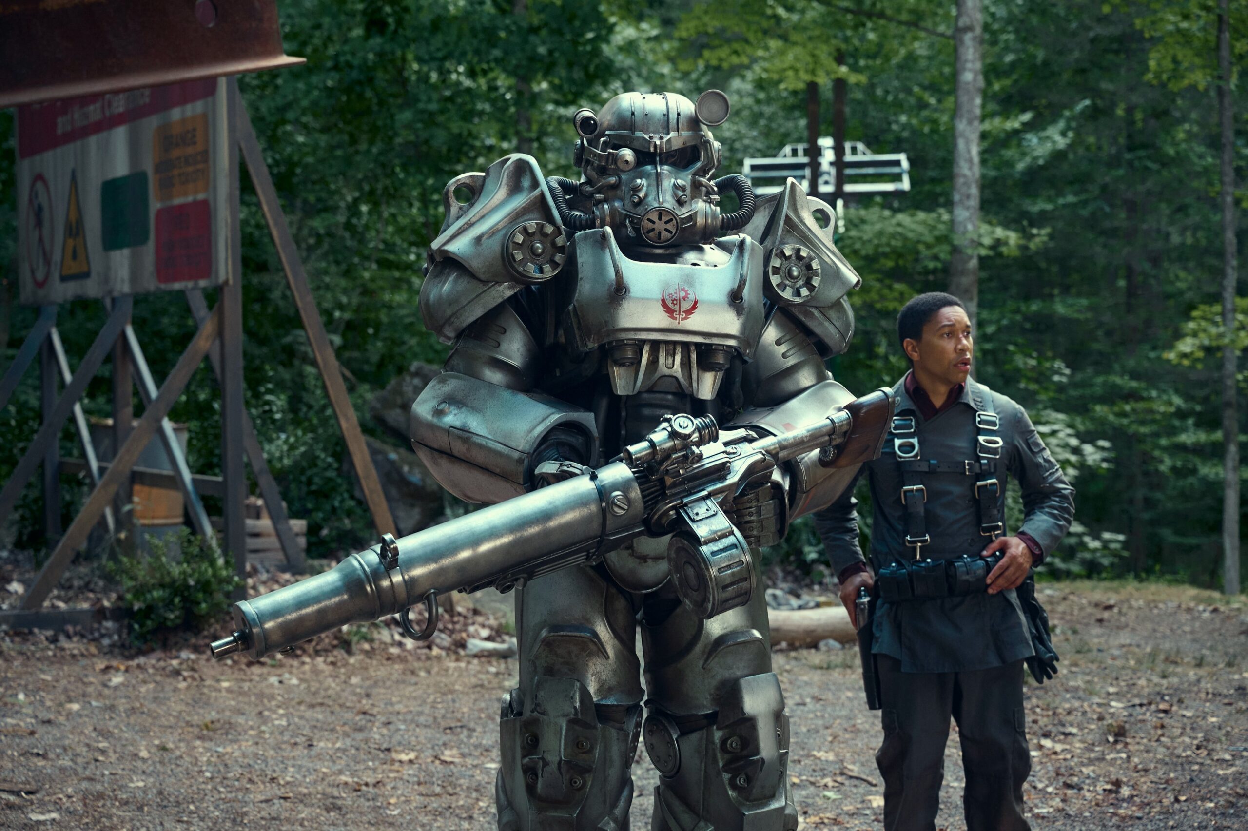 Postać w futurystycznym pancerzu z przypominającym minigun uzbrojeniem stoi obok mężczyzny w taktycznym ubiorze, w tle leśne otoczenie.