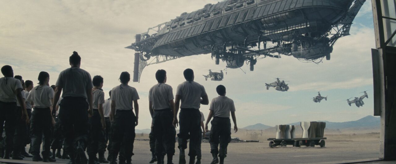 Grupa ludzi w mundurach obserwuje olbrzymi statek kosmiczny i latające pojazdy przypominające helikoptery w hangarze lotniczym.