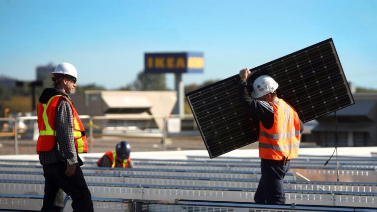 Pracownicy w kaskach i kamizelkach odblaskowych instalują panele słoneczne na dachu, w tle widoczny napis IKEA.