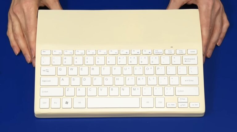 Zdjęcie kremowej klawiatury Raspberry Pi 500 znajdującego się na niebieskim tle, obsługiwanej przez dwie ludzkie dłonie.