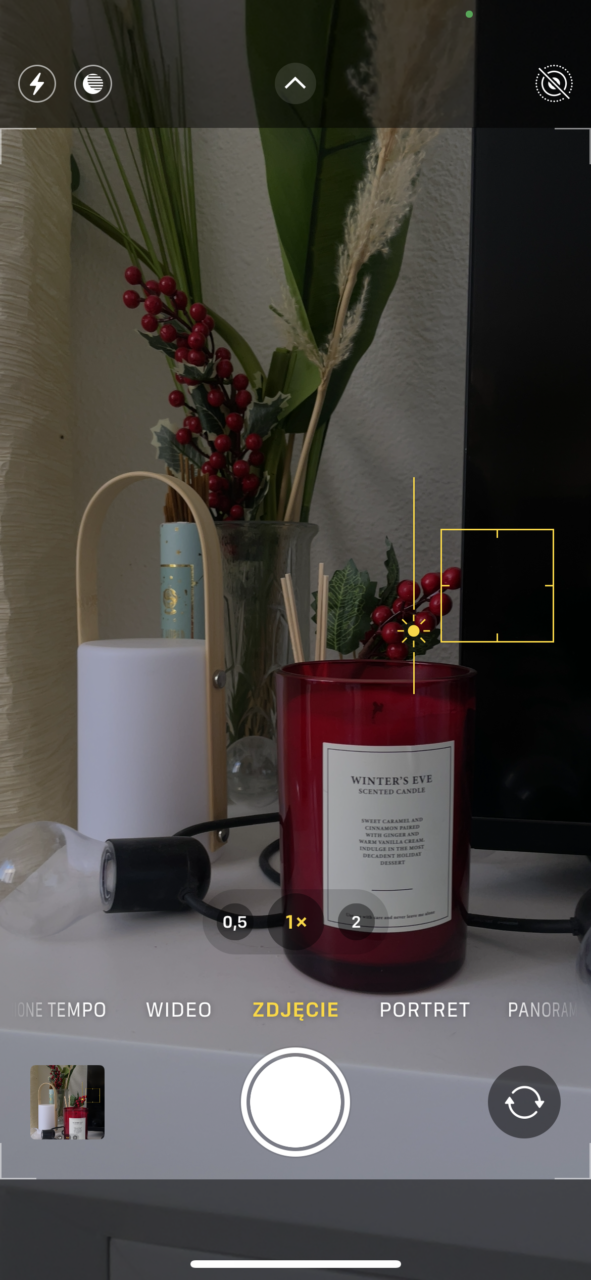 Zrzut ekranu iphone aparatu telefonu, skupiający się na zapalonej czerwonej świecy zapachowej, obok której stoi biała lampa i znajdują się dekoracyjne gałązki z czerwonymi owocami. Na dole ekranu widać kontrolki aparatu.