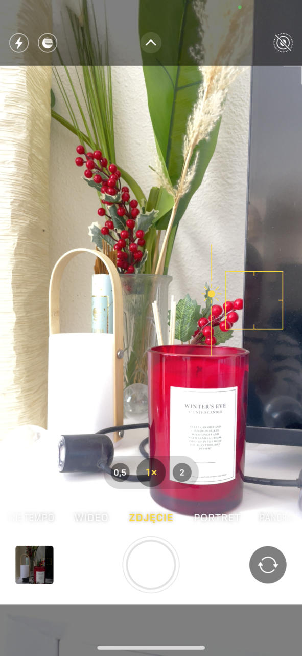 Zrzut ekranu iphone aparatu telefonu, skupiający się na zapalonej czerwonej świecy zapachowej, obok której stoi biała lampa i znajdują się dekoracyjne gałązki z czerwonymi owocami. Na dole ekranu widać kontrolki aparatu.