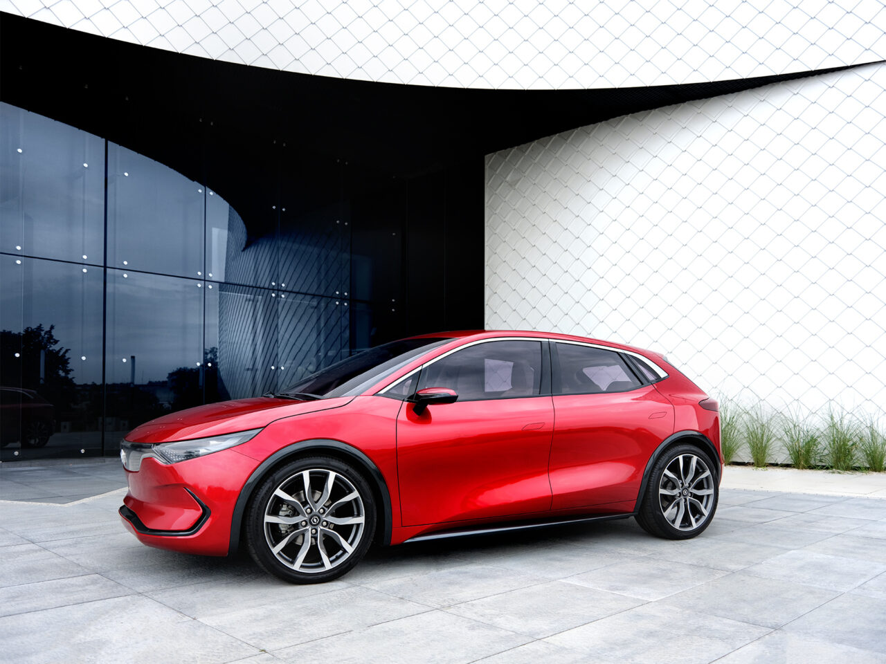 Czerwony samochód elektryczny Izera zaparkowany przed budynkiem ze szklaną fasadą i białymi, geometrycznymi elementami.